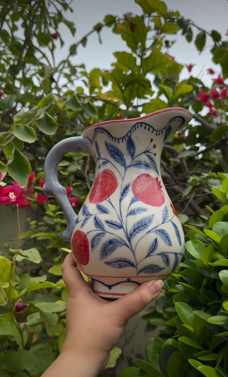 Hand holding a hand-painted jug with red pomegranates and blue leaves against flowering greenery